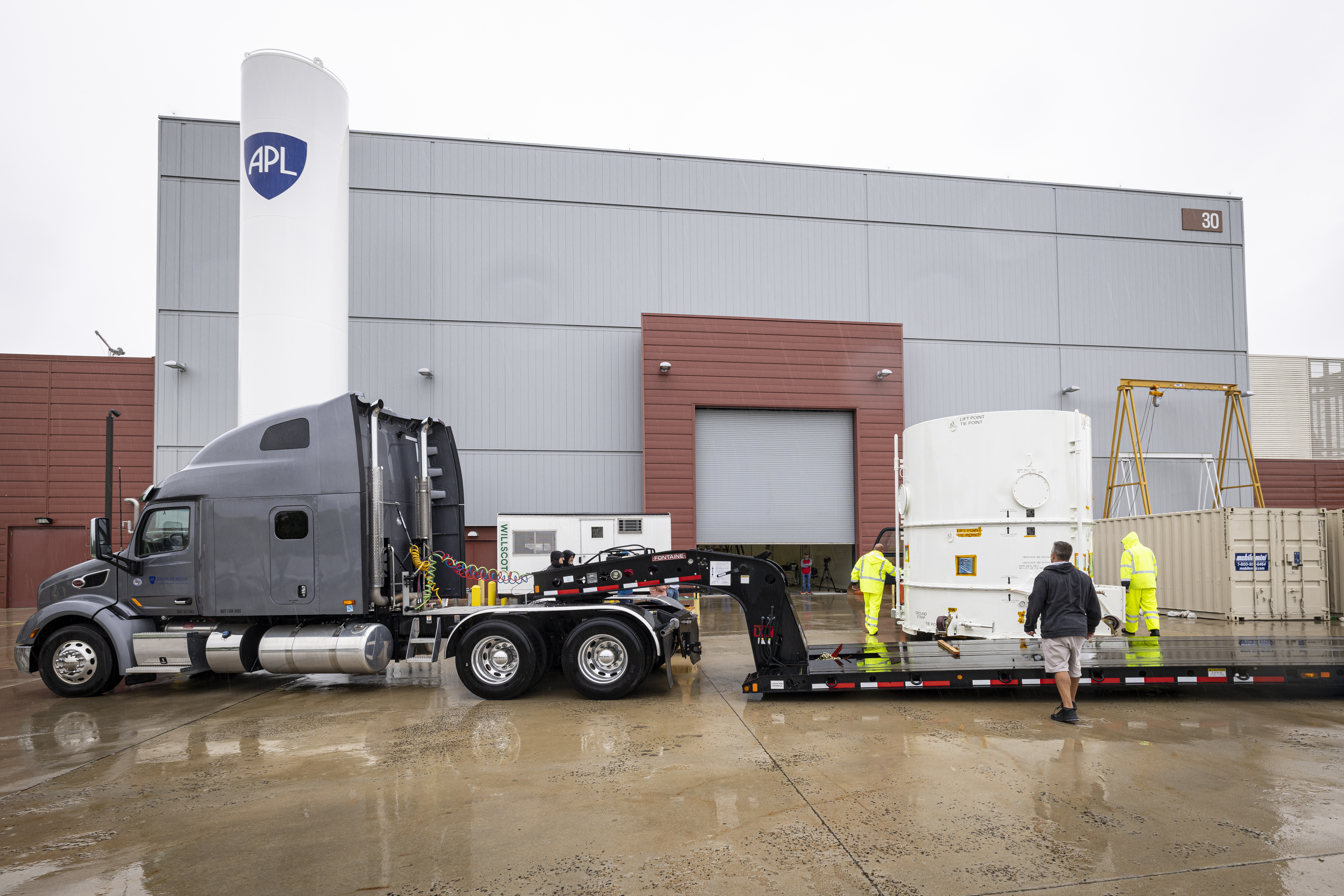A tractor trailer sits parked outside a building with the APL shield on it and a white container with the Europa Clipper modules