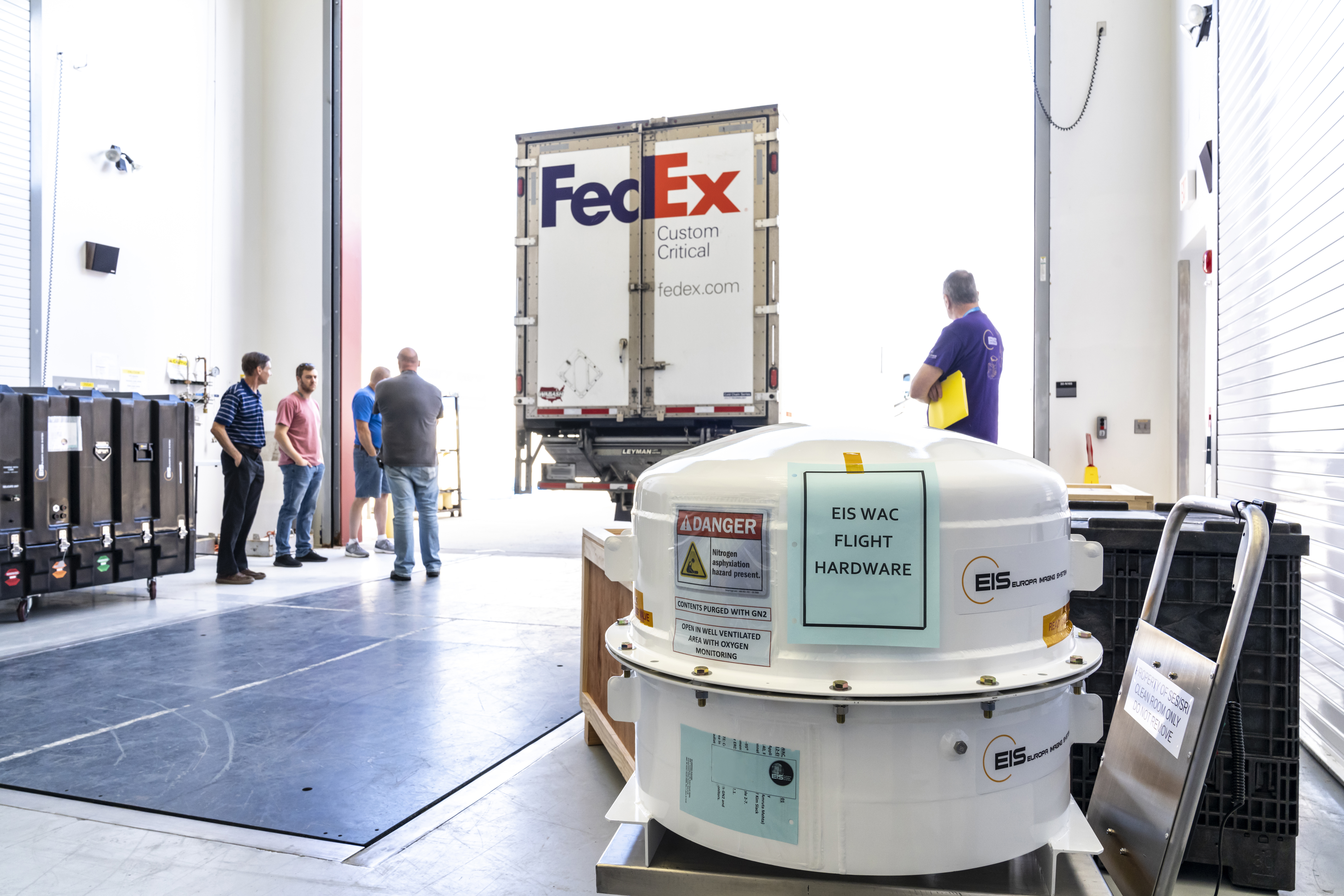 A white container with the a paper reading "EIS WAC Flight Hardware" sits in a room with three people and a FedEx truck