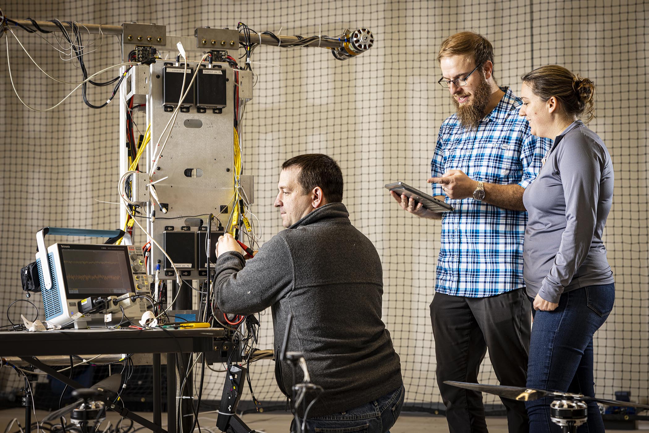 Three researchers stand in front of a test platform of the Dragonfly electronics, one kneeling while looking at a voltmeter, another holding a tablet