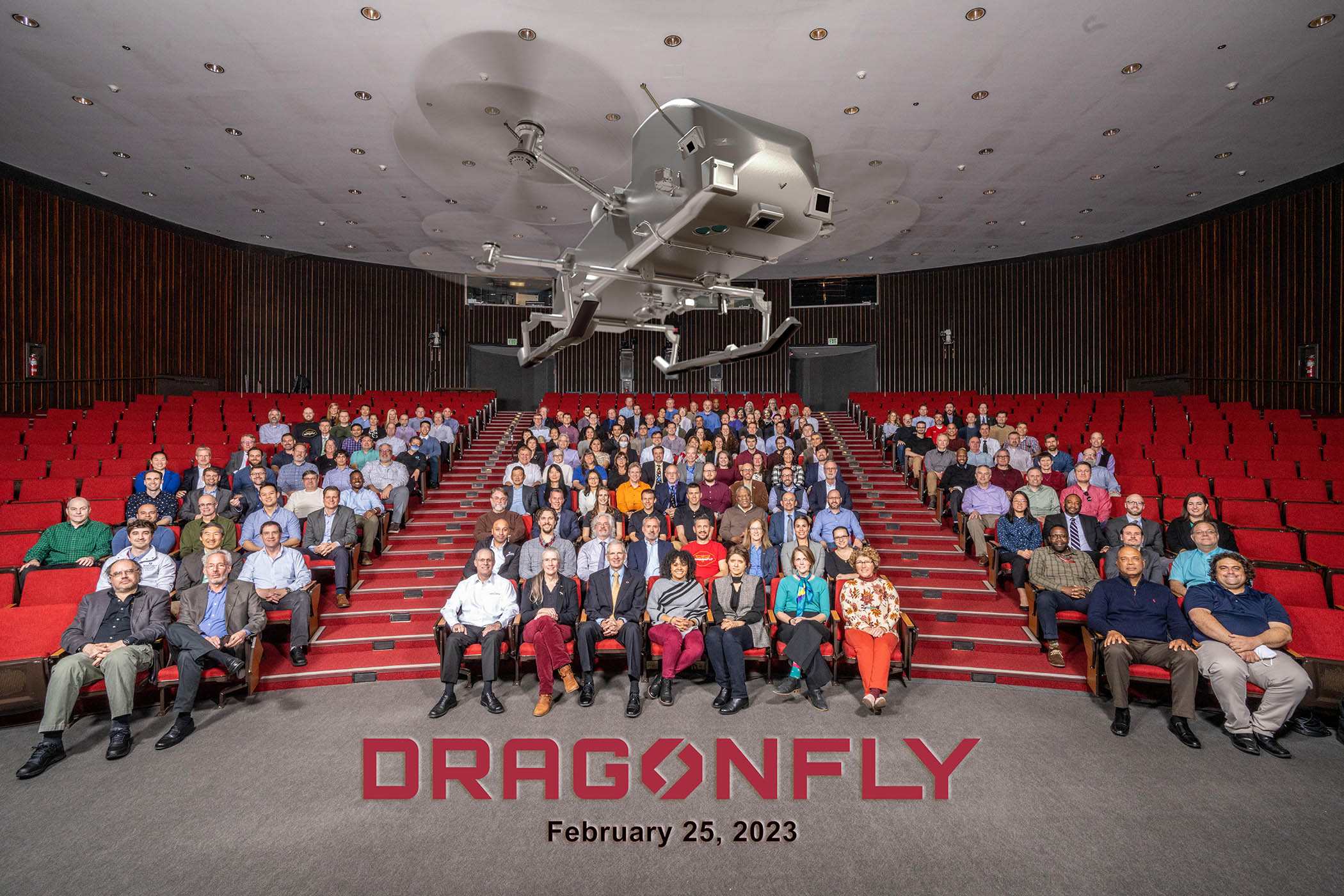 A group of people sit in an auditorium full of red chairs while an illustration of the Dragonfly rotorcraft flies above them