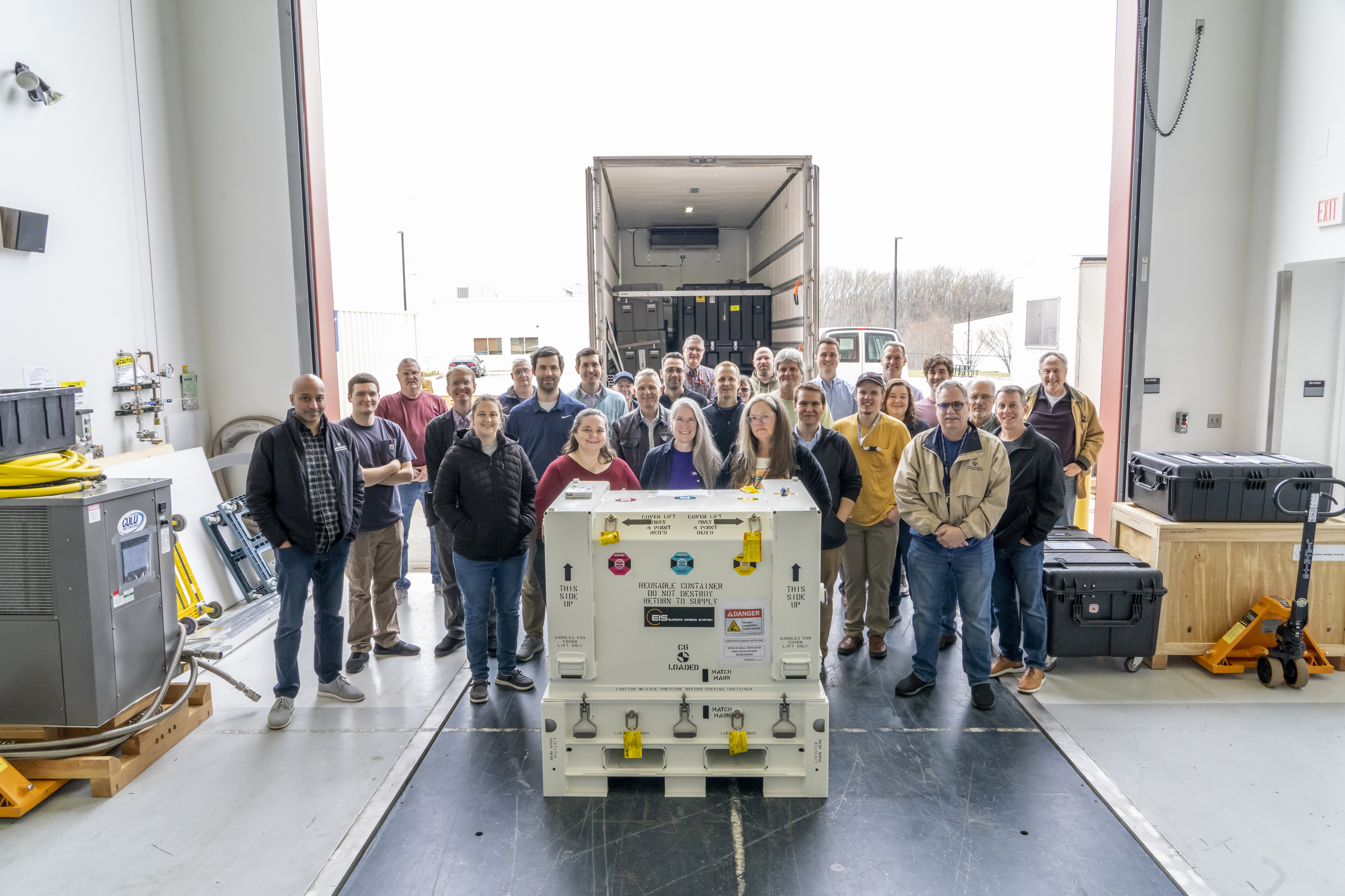 A group of people stand in front of the open doors of a FedEx truck and behind a closed white temperature-sensitive box that&#039;s holding the EIS NAC instrument