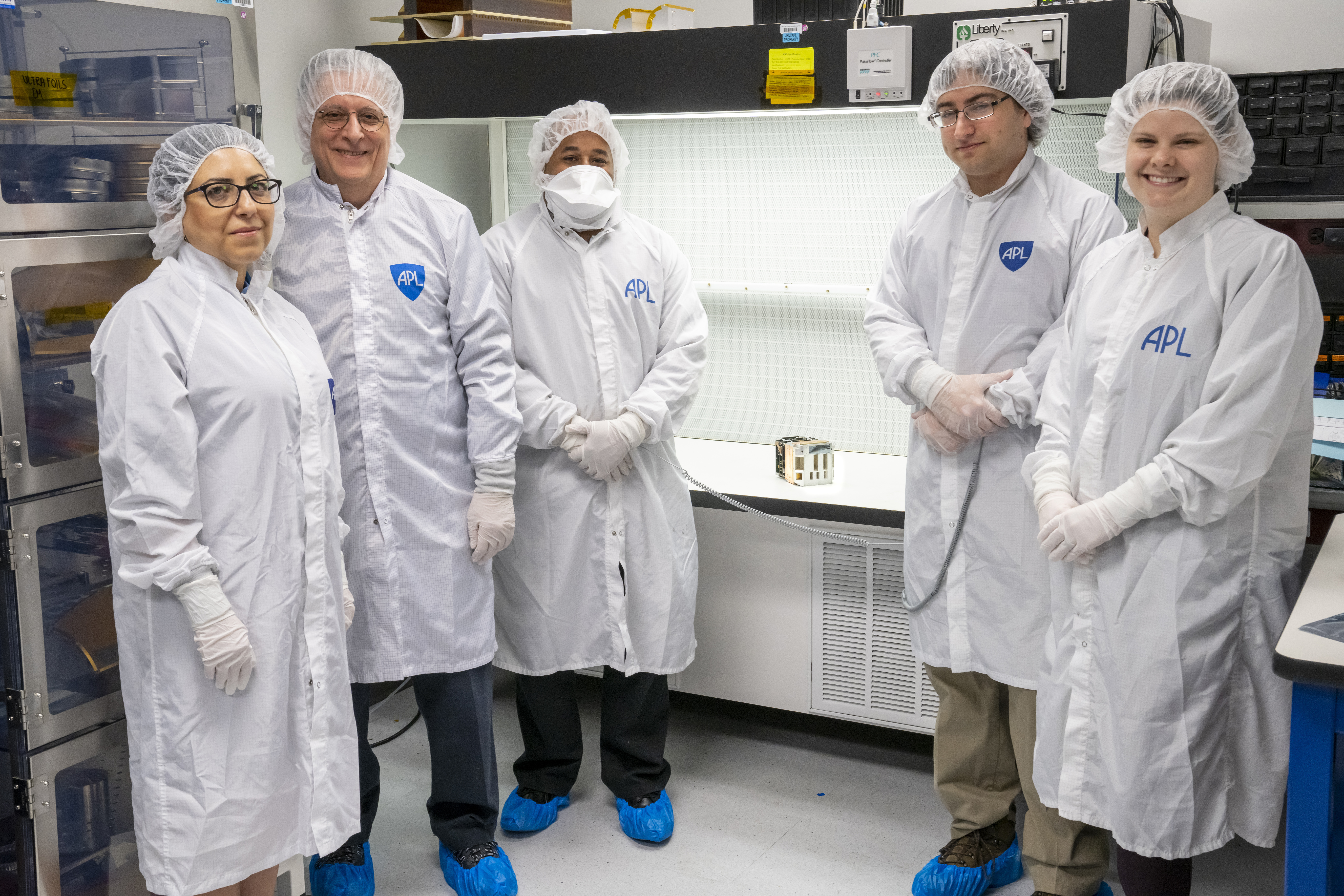 Five people wearing white hair nets, white masks, white gloves and white lab coats with the APL logo stand around a lab table with an electronics box on it