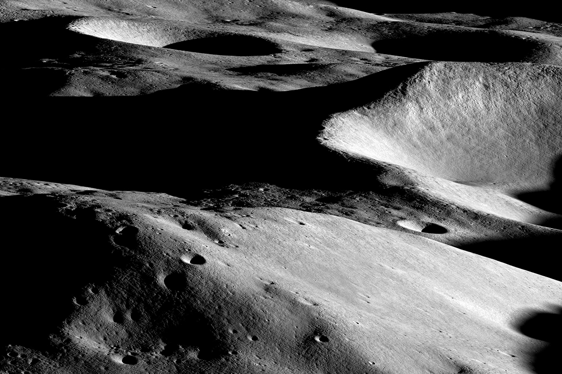 An image show partly side-lit, partly shadowed, gray hills on the Moon, with both large and small craters covering the surface