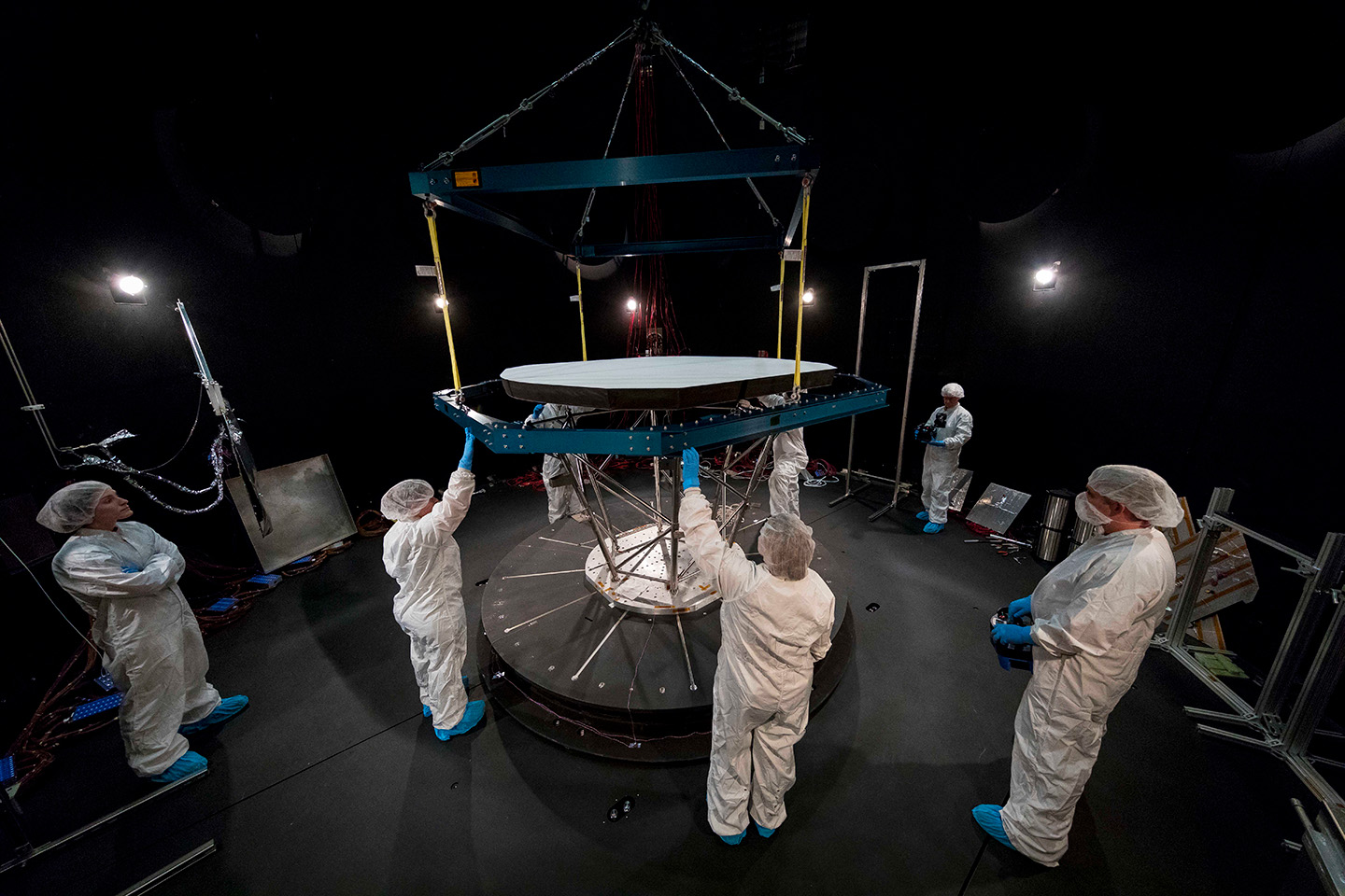 Seven people dressed in white bunny suits, hair nets, booties, and blue latex gloves stand in a dark room with lights shining on the Parker Solar Probe heat shield above them