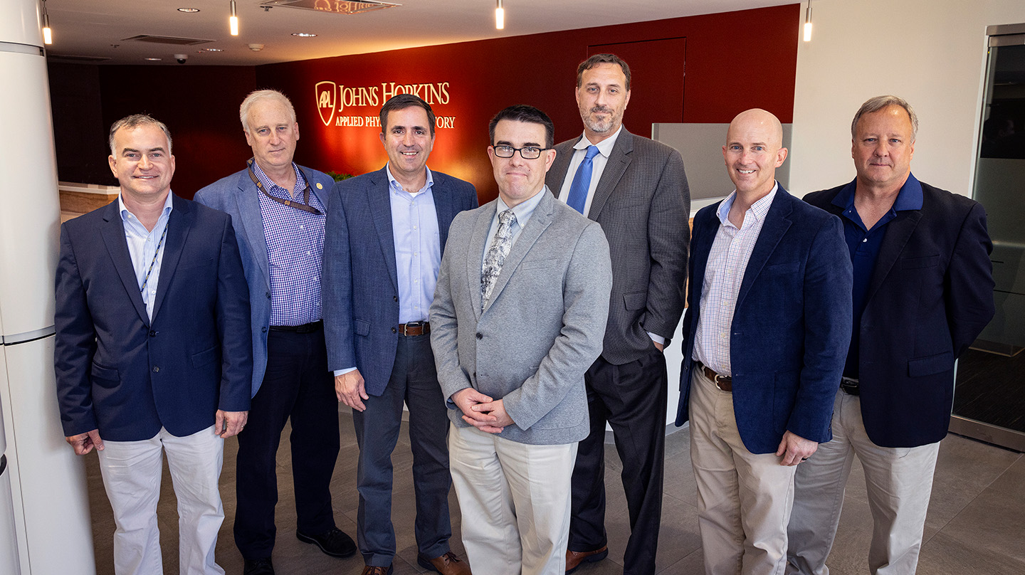 A group of men wearing suits stand together in a foyer with the logo and title of the Johns Hopkins Applied Physics Laboratory behind them on a red wall