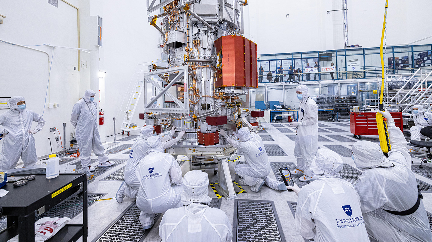 People wearing white bunny suits, hairnets, masks and booties inspect the Europa Clipper propulsion module in a white clean room