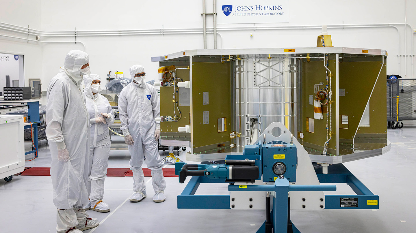 Two people wearing white bunny suits, booties, and head coverings examine the metallic scaffold of the IMAP spacecraft