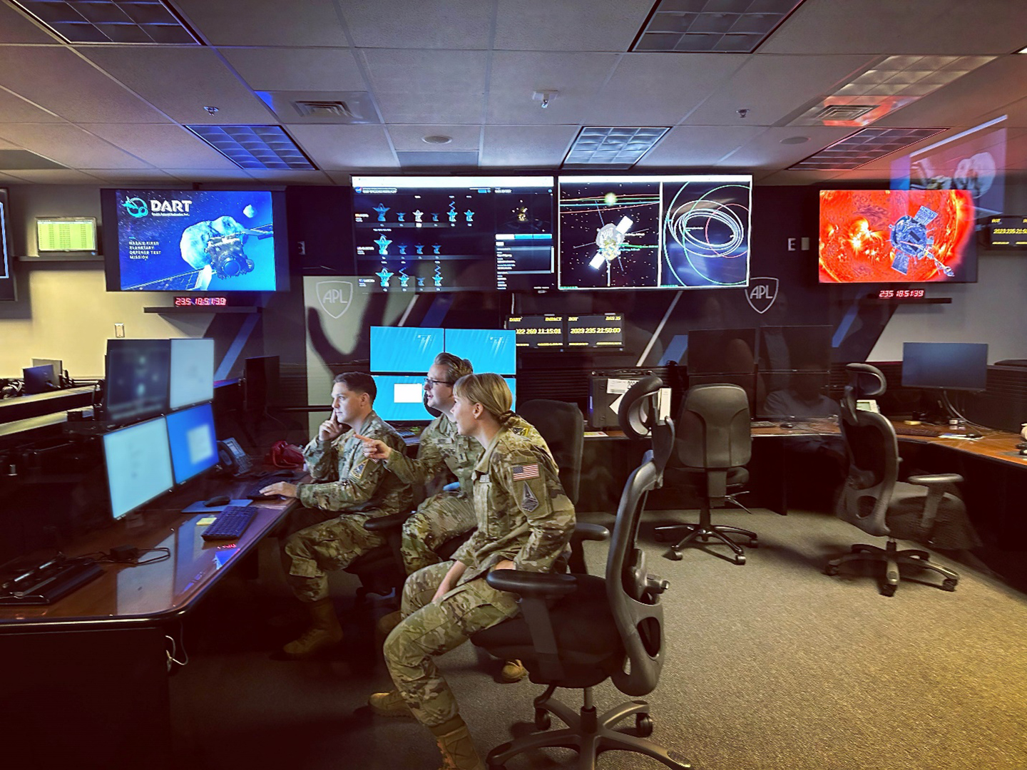 Three people wearing military camouflage uniforms sit at a desk and computer monitor. Several screens behind them list other NASA missions, including DART and Parker Solar Probe