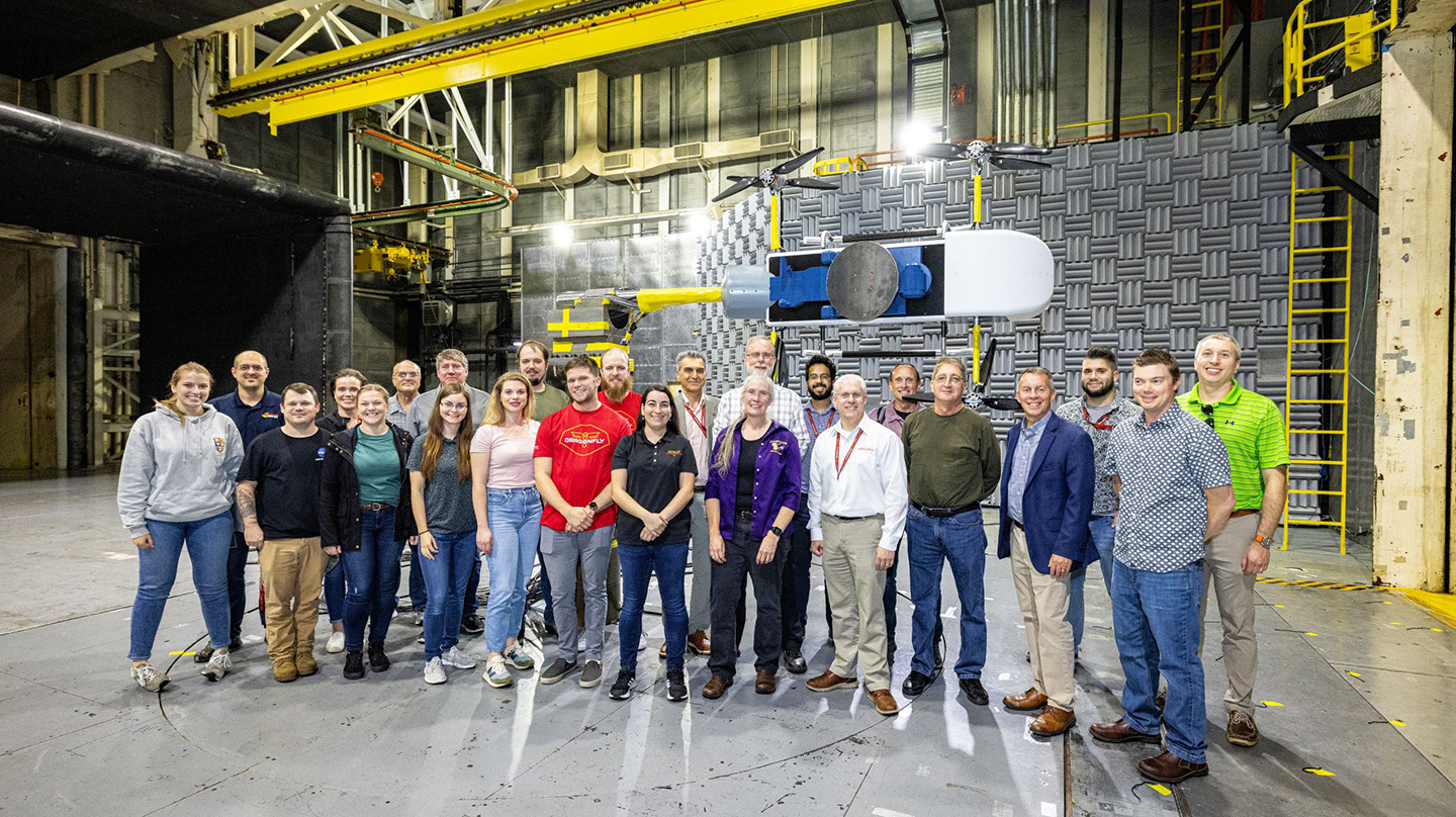 A group of people stand together for a photo in a large metallic test room with a model of the Dragonfly spacecraft hanging behind them