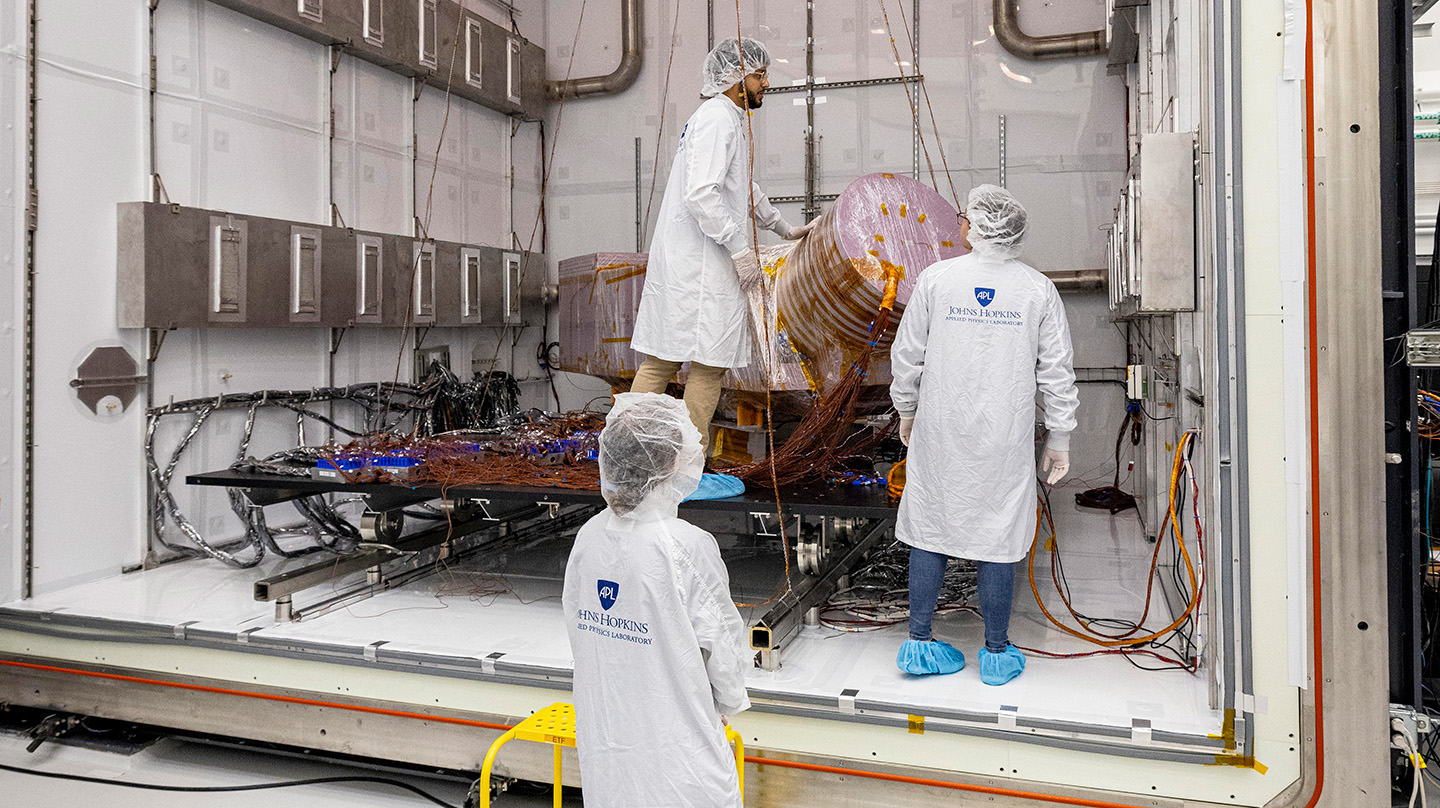 Three people wearing white lab coast, white hair nets and blue booties work on assembling and wiring a model of the Dragonfly spacecraft while in a giant white box