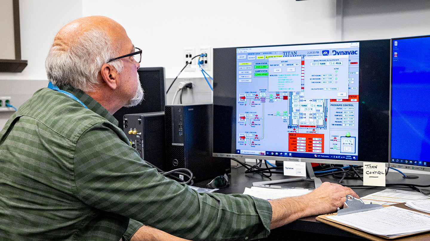 A partly bald man wearing glasses and a green checkered collared shirt looks at a computer monitor where measurements from the Titan Chamber are visible