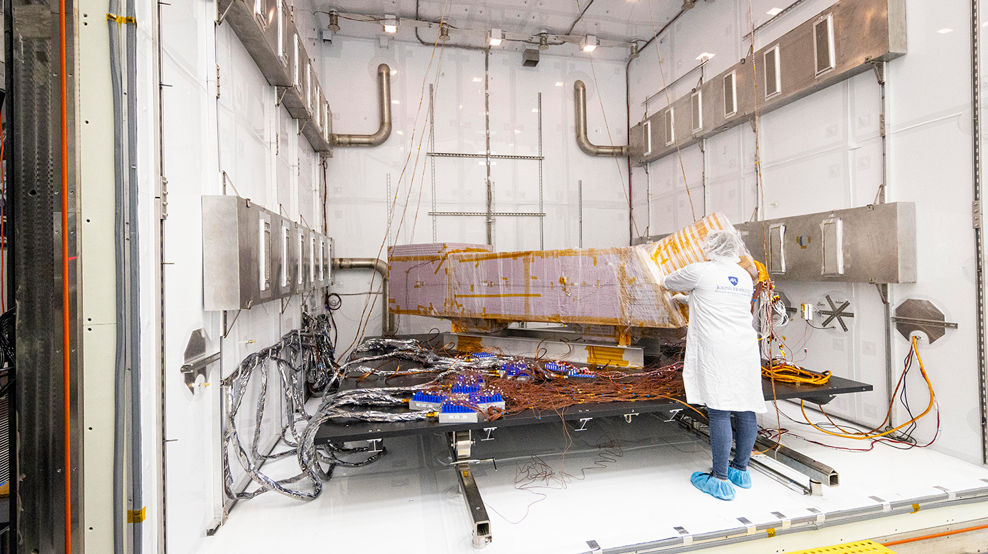 A person wearing jeans, blue booties, a white lab coat and a white hair net stands inside a large white box with various pipes and boxes distributed throughout, all while working on a model of the Dragonfly spacecraft