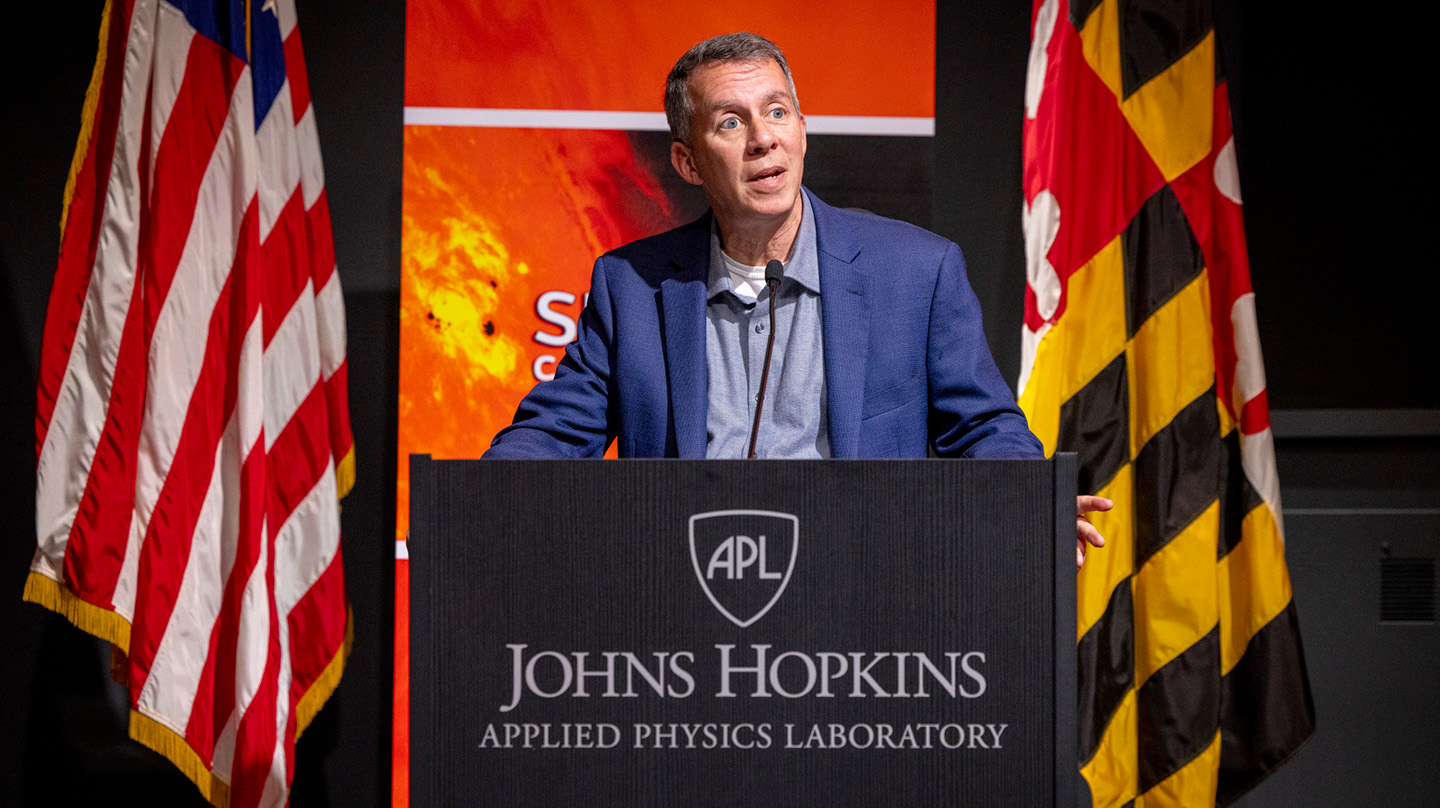 A photo of Bobby Braun standing at a podium between the American and Maryland flags, with a placard in front that says &quot;Johns Hopkins Applied Physics Laboratory&quot;