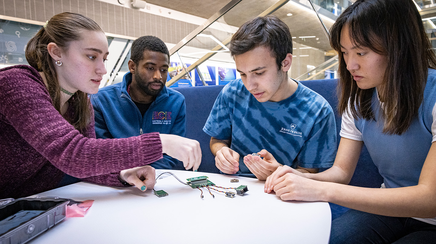 Three students sit with a mentor at table where they work on putting together electronics chips and wires