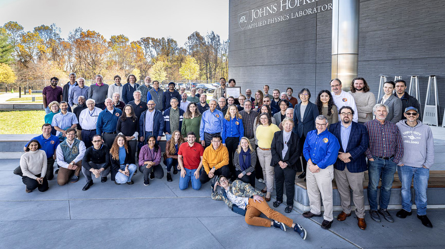 A photo of the Parker Solar Probe team, standing outside under an awning, with fall leaves in the background and a sign reading, "Johns Hopkins Applied Physics Laboratory"
