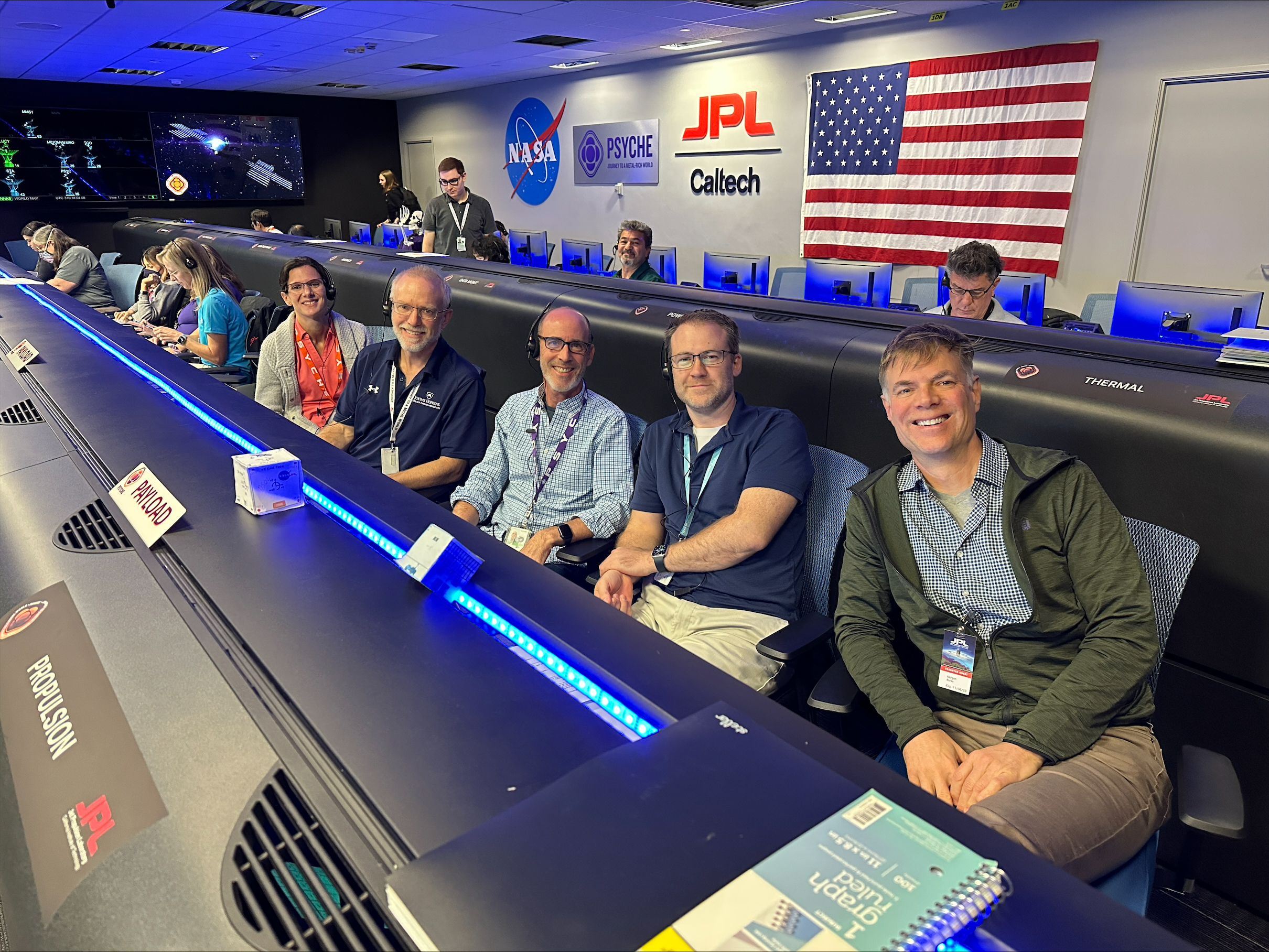 Several people sit behind a table with a blue neon light and an American flag, the NASA logo, the JPL logo and the Psyche mission logo are on the wall behind them