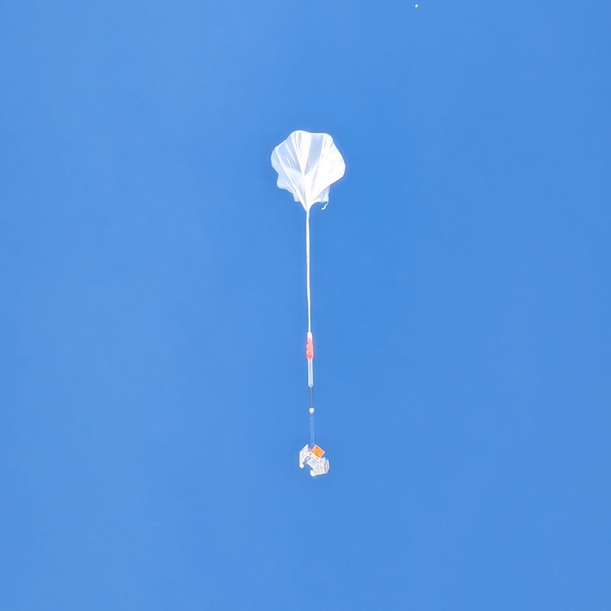A white balloon floats into the blue sky, a gondola and telescope dangling far below it