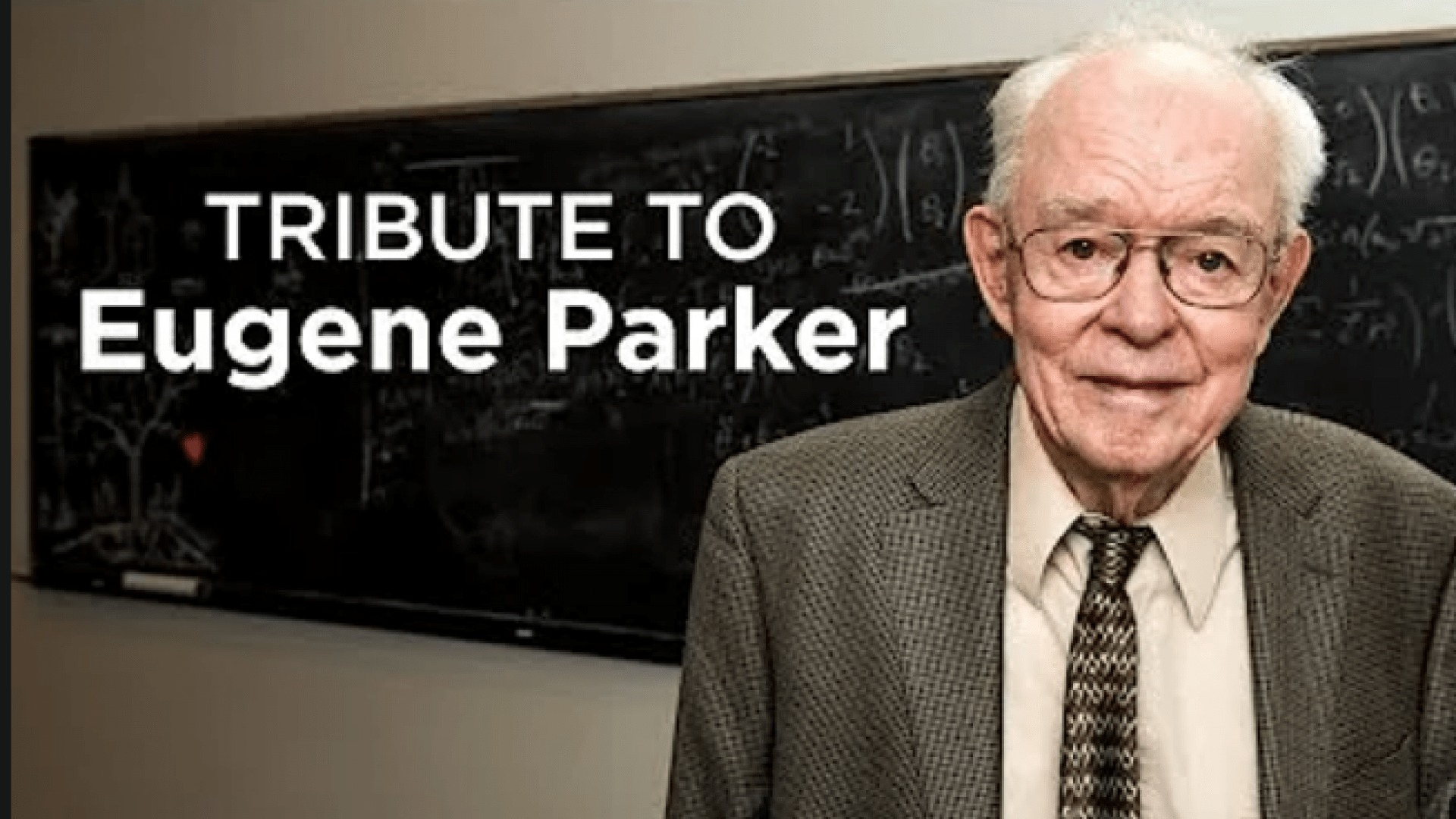 Eugene Parker stands to right with the words &quot;Tribute to Eugene Parker&quot; over his right shoulder. He&#039;s standing in front of a chalkboard with math equations written on it.