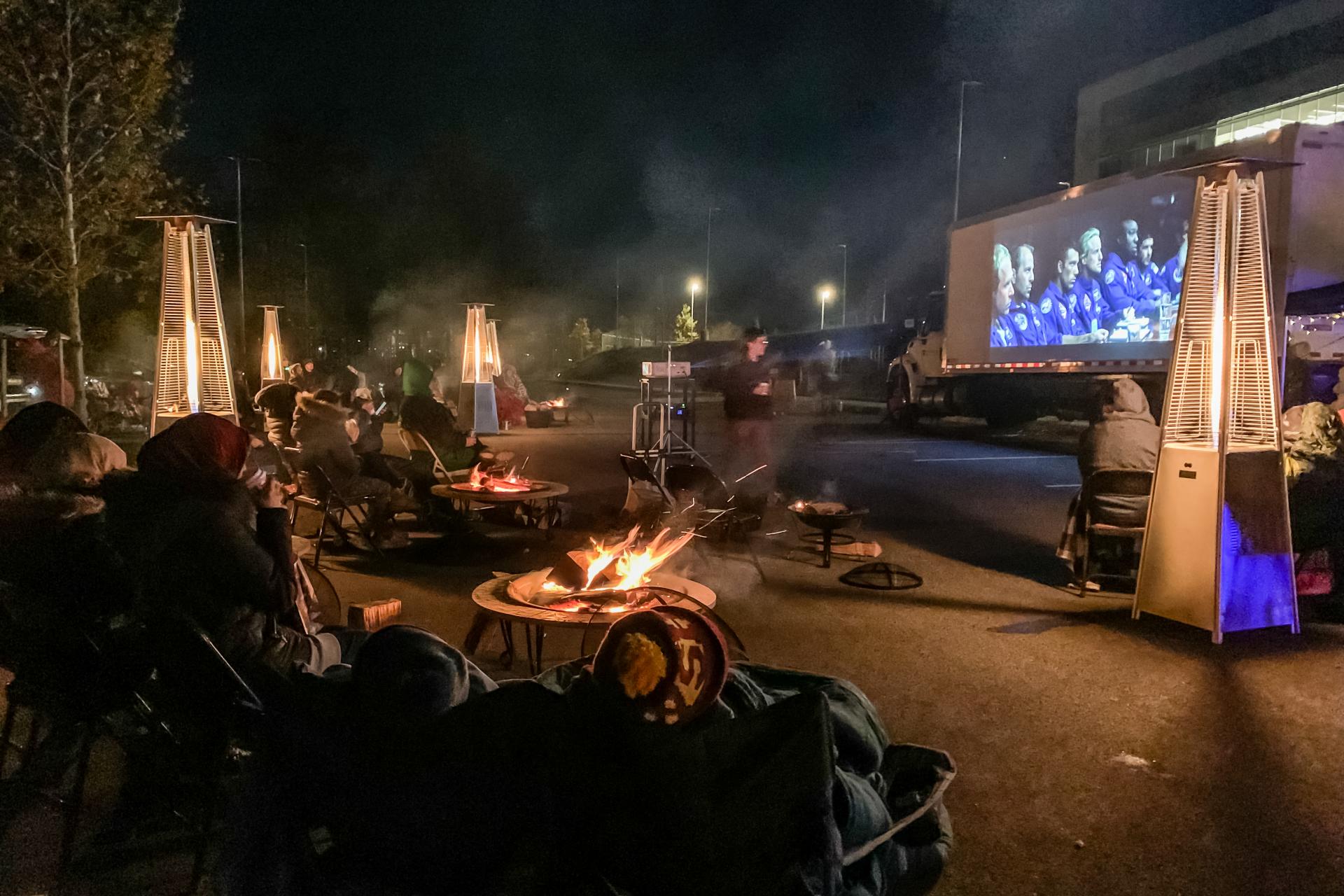 People sit outside wrapped in blankets watching a movie with heaters around them