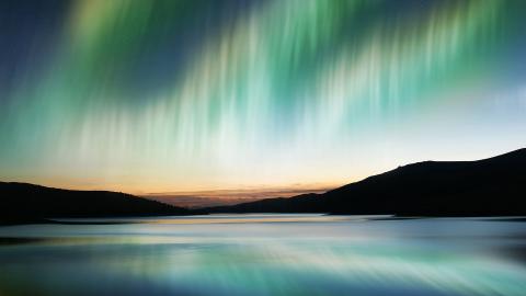 A blue and green aurora appearing over a body of water and a mountain range in the background
