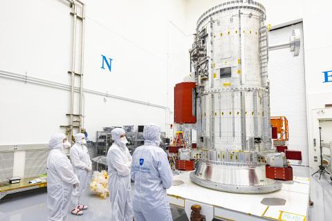 People wearing bunny suits in a cleanroom look up at a massive metal cylinder, the Europa Clipper propulsion module