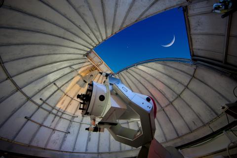 A telescope observatory with a ceiling window open to the blue sky and crescent Moon above