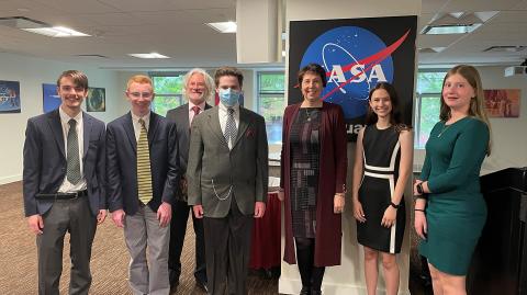 Seven people dressed in formal attire stand together with a NASA logo behind them