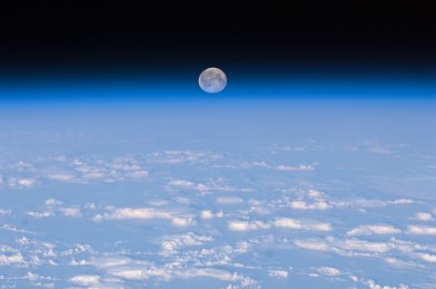 A photo from space shows the Moon rising over the curvature of the blue Earth. Clouds in the sky show at the foreground of the picture