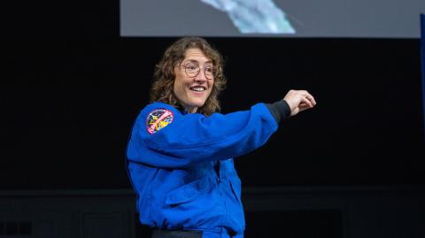 Christina Koch, wearing her NASA astronaut jacket, lifts her arm while giving her colloquium to APL staff 