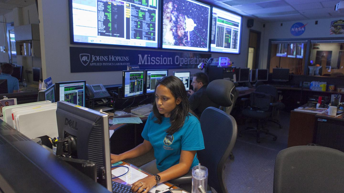 Woman at a computer desk that is part of the large mission operations room with three large projection screens on the back wall