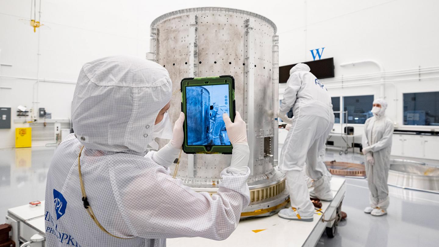 Three APL engineers in white clean room suits, one taking a photo with an iPad of another working on a large metal tank